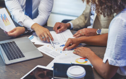 Close up of people pointing to a graph with data. Close up of people pointing to a graph with data. There is also a digital tablet and a laptop on the table.